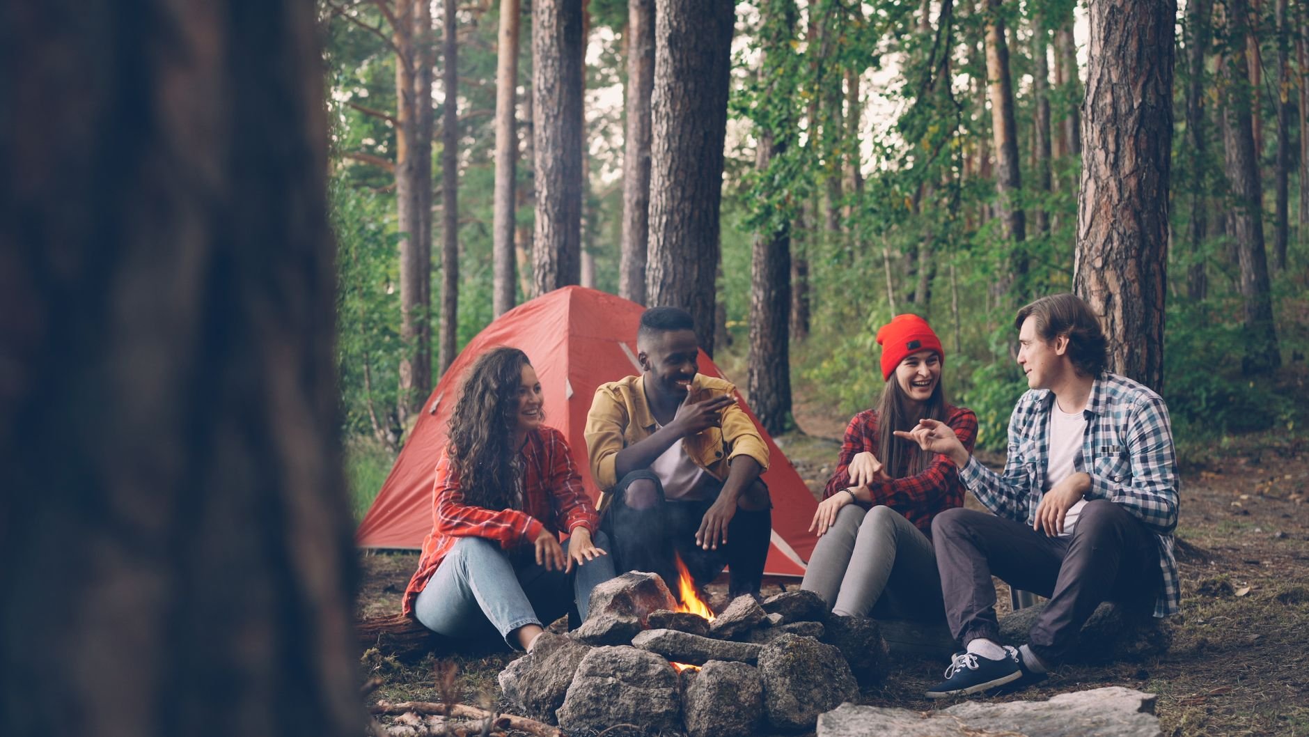 A group of friends gathered around a campfire in a forest next to a tent.